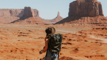 Eine Frau mit Rucksack und einem Hut in einer Sand Wüste mit großen Felsen   