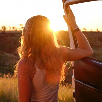 Frau lehnt an ihrem Auto und blickt auf einsame Landschaft bei Sonnenuntergang
