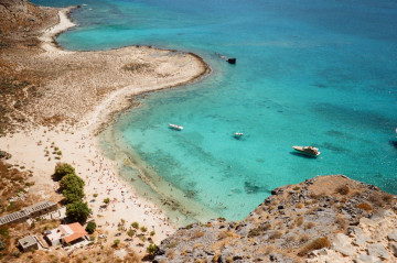 Sandstrand mit Menschen und einem Kleinen Haus Links unten im Bild und ein drei Booten auf dem Meer auf Kreta