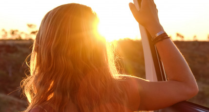 Blonde Frau blickt, angelehnt an ihr Auto, auf einsame Landschaft bei Sonnenaufgang
