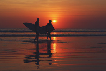 Zwei Menschen die mit einem Surfbrett am Strand bei Sonnenuntergang spazieren gehen 